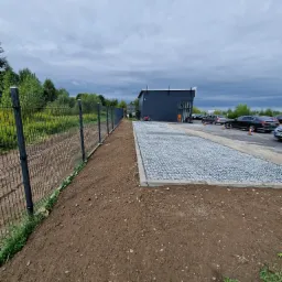 Ukończona nawierzchnia z jasnych kamieni na parkingu przed budynkiem z szarą elewacją, widoczny fragment ogrodzenia z siatki, przygotowany teren pod nasadzenia i zaparkowane samochody.
