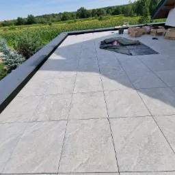 Jasnoszare płytki ułożone na płaskim dachu budynku jednorodzinnego, widoczne kartony i materiały budowlane, w tle zieleń drzew i pola.