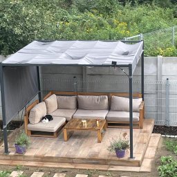 MC-BRUK Michał Chmielewski - Zadaszony taras z drewnianą podłogą wyłożoną płytkami imitującymi deski, na którym stoi narożna sofa z jasnymi poduszkami i drewniany stolik. Na sofie leży czarno-biały kot. W tle widoczny ogród...