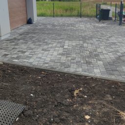 MC-BRUK Michał Chmielewski - Świeżo ułożona, szara kostka brukowa przed budynkiem z brązową bramą garażową, widoczny fragment przygotowanego terenu pod dalsze prace.