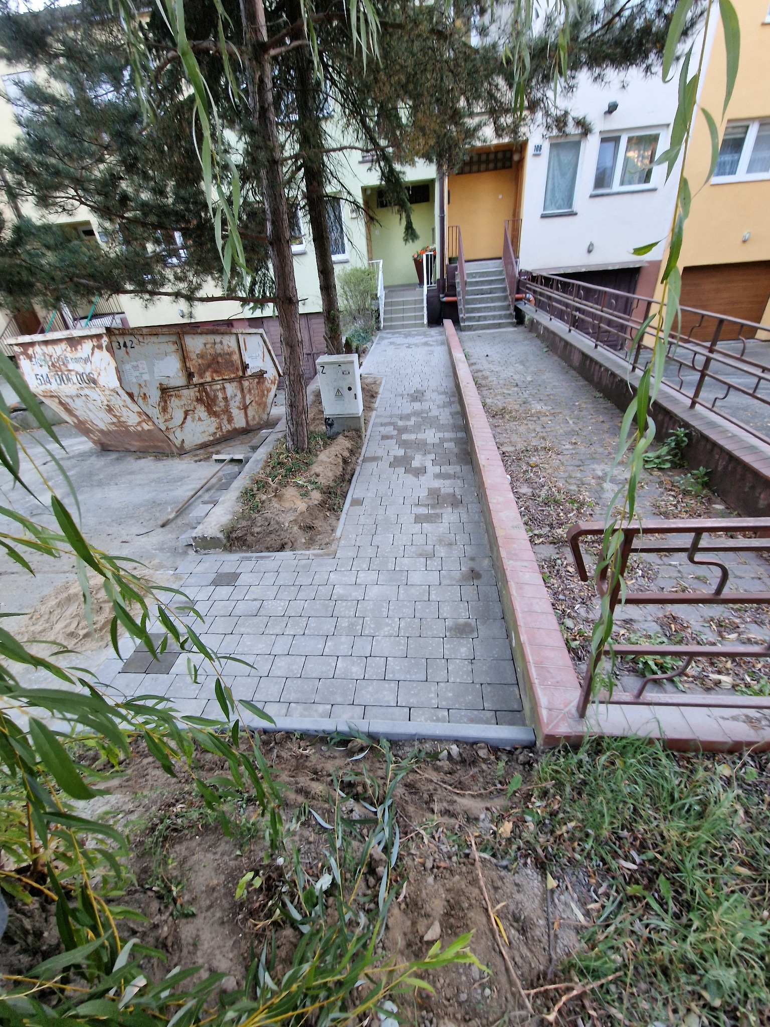 Świeżo ułożona, szara kostka brukowa prowadząca do budynku mieszkalnego, z widocznym kontenerem na gruz w tle i młodymi drzewkami wzdłuż ścieżki.