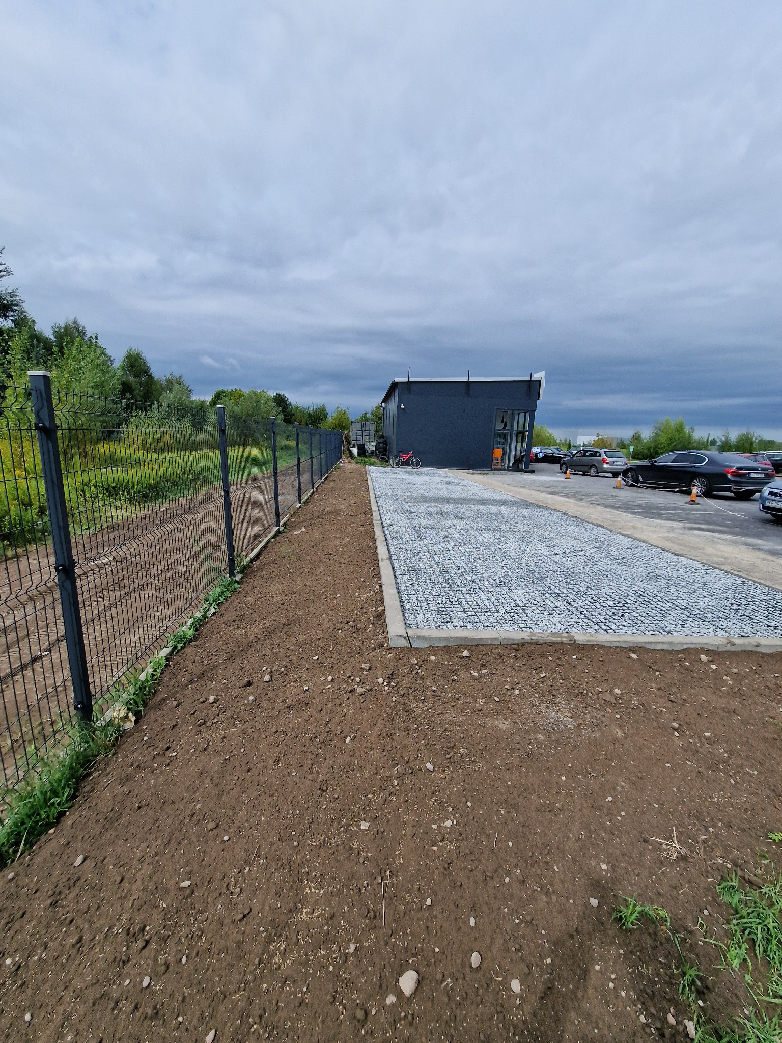 Ukończona nawierzchnia z jasnych kamieni na parkingu przed budynkiem z szarą elewacją, widoczny fragment ogrodzenia z siatki, przygotowany teren pod nasadzenia i zaparkowane samochody.