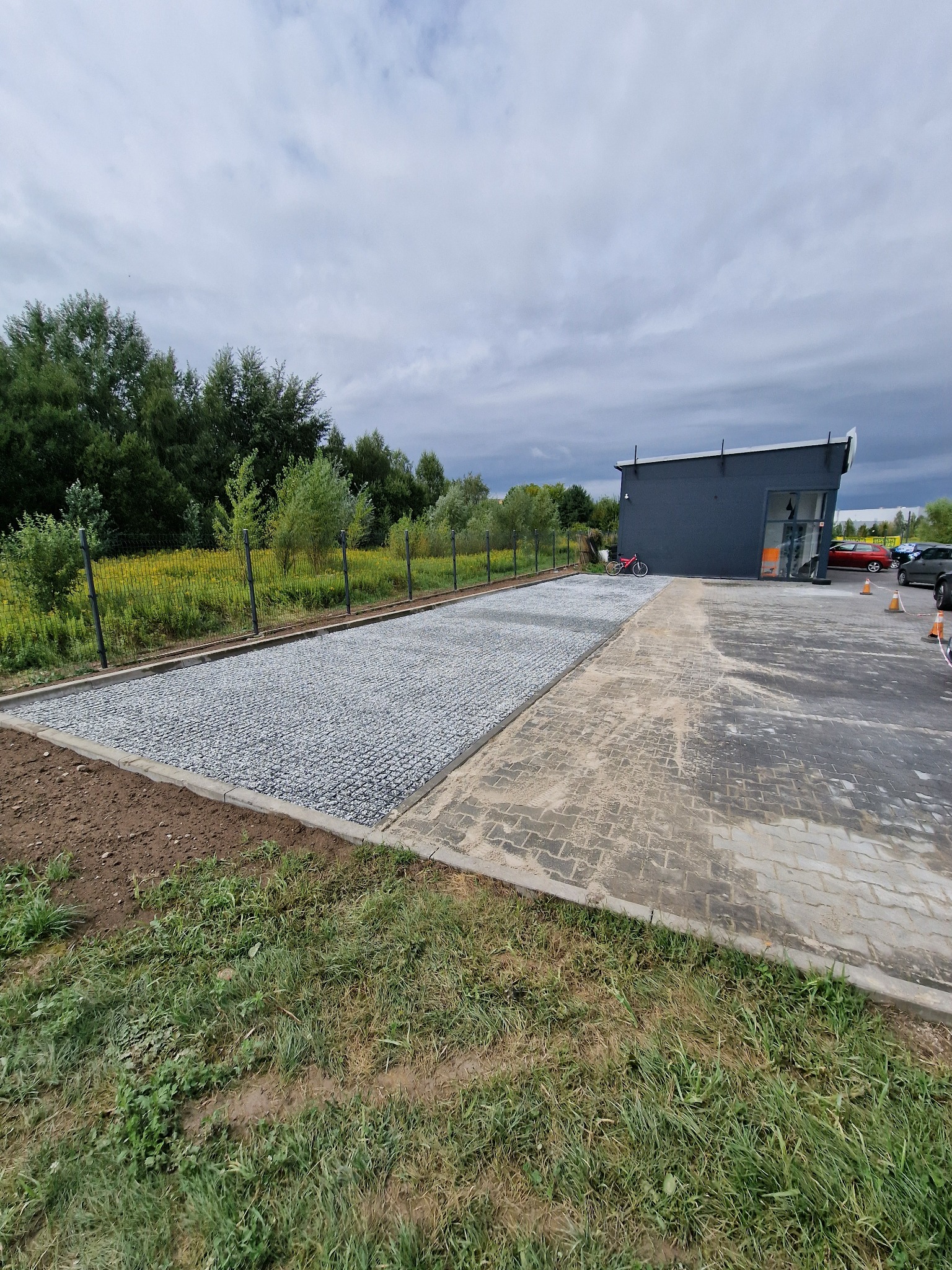Nowo utwardzona powierzchnia z drobnego kruszywa obok parkingu z kostki brukowej, budynek w tle, ogrodzenie i zieleń.