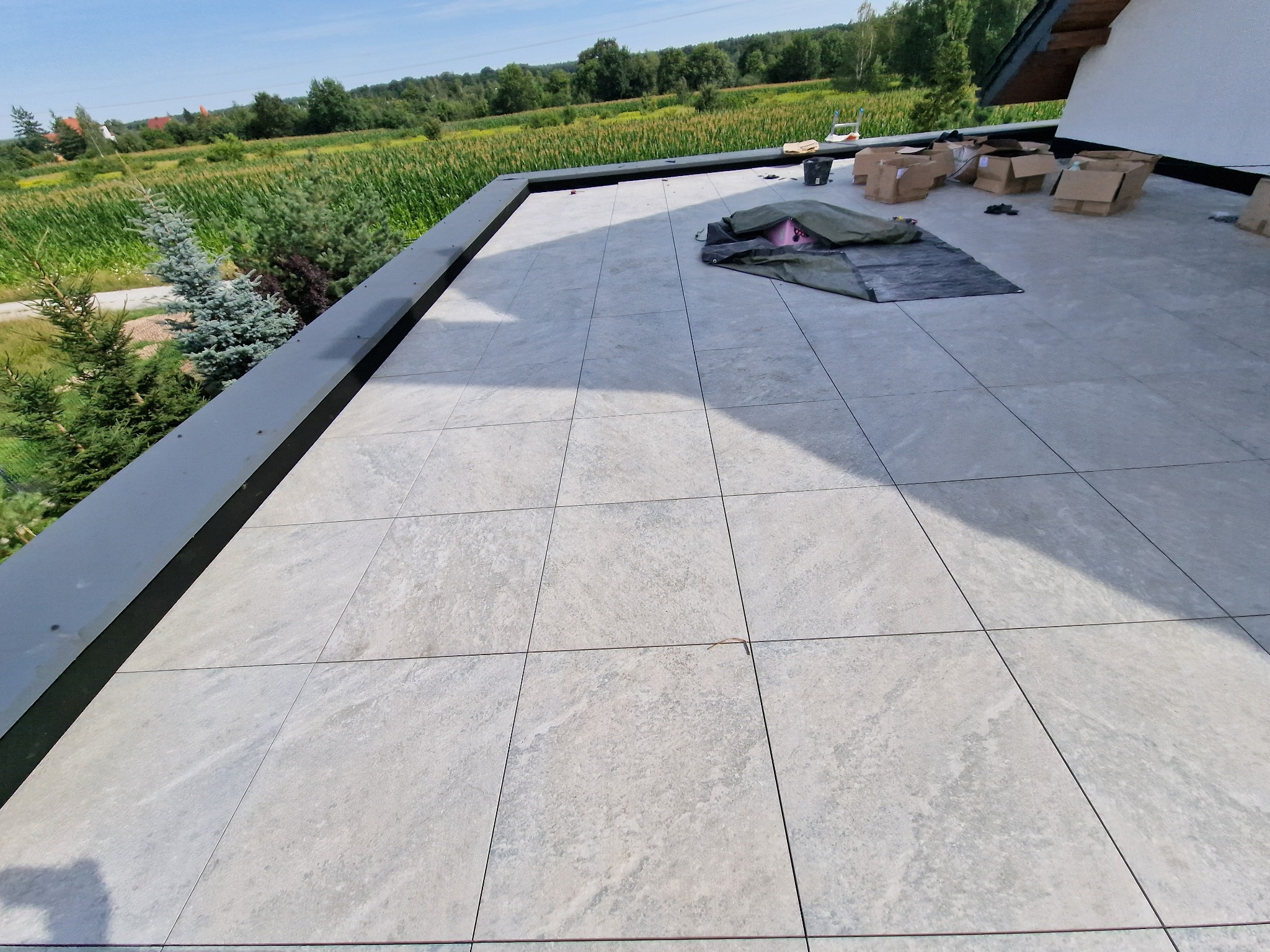 Jasnoszare płytki ułożone na płaskim dachu budynku jednorodzinnego, widoczne kartony i materiały budowlane, w tle zieleń drzew i pola.