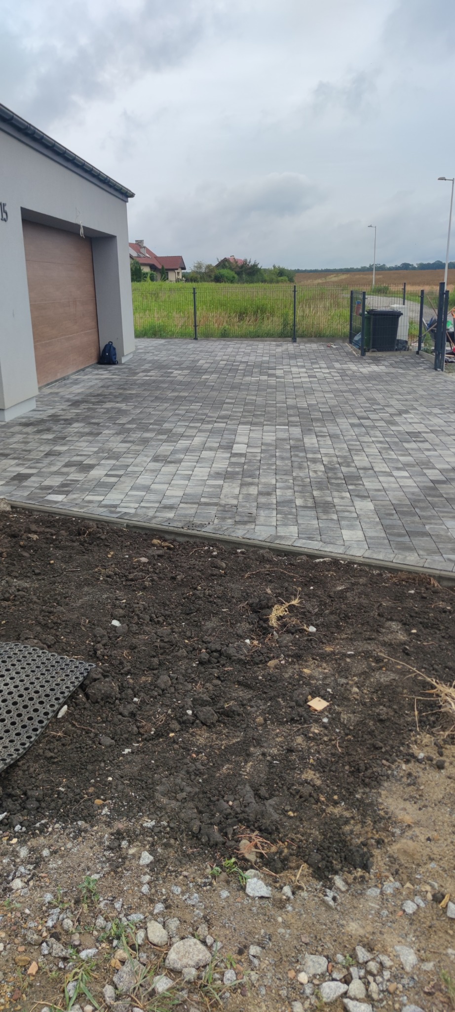 Świeżo ułożona, szara kostka brukowa przed budynkiem z brązową bramą garażową, widoczny fragment przygotowanego terenu pod dalsze prace.