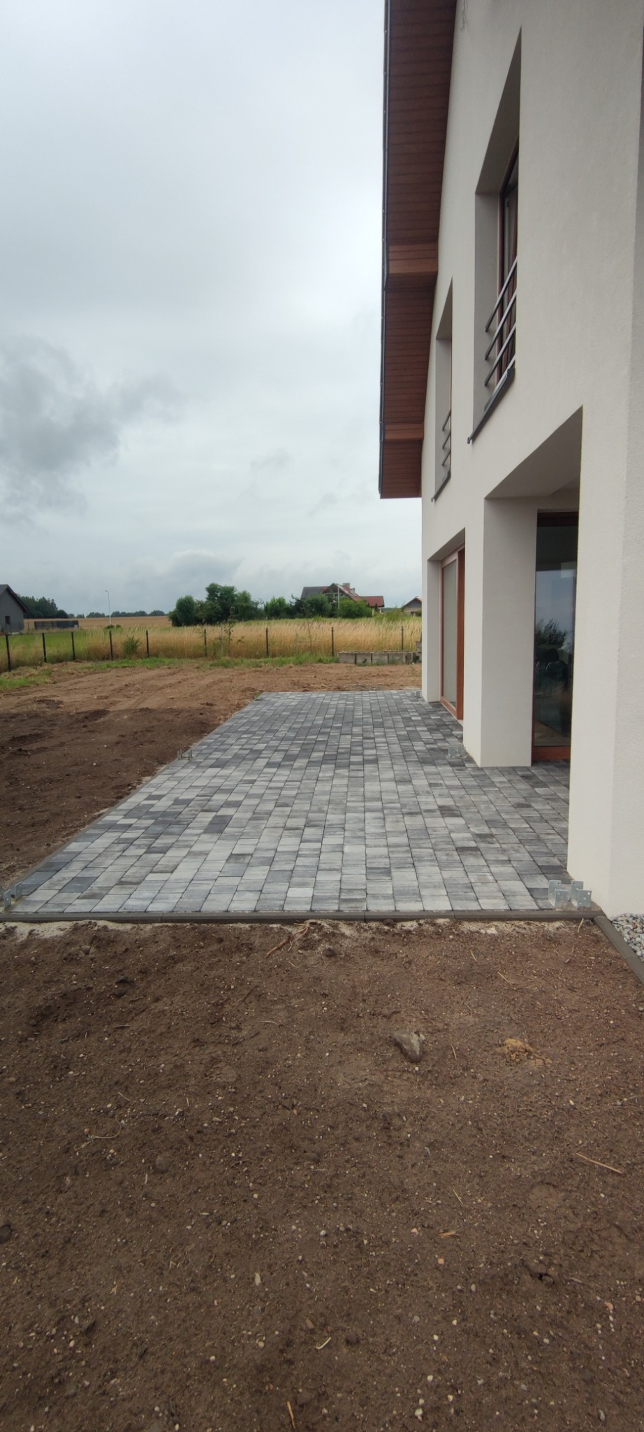 Nowo ułożony taras z szarej kostki brukowej przy białym domu jednorodzinnym, widoczna ziemia przygotowana pod trawnik i ogrodzenie w tle.