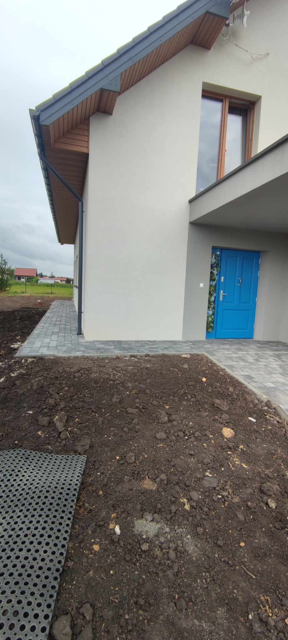 Ułożona kostka brukowa prowadząca do niebieskich drzwi wejściowych nowoczesnego domu, z widoczną przygotowaną ziemią pod dalsze prace brukarskie i czarną matą drenażową.
