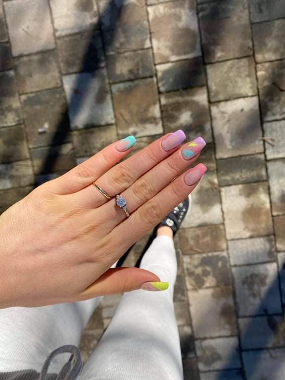 Dłoń z pierścionkiem zaręczynowym prezentuje geometryczny manicure: pastelowe końcówki w odcieniach turkusu, fioletu, różu i limonki, na tle szarej kostki brukowej i białych spodni.