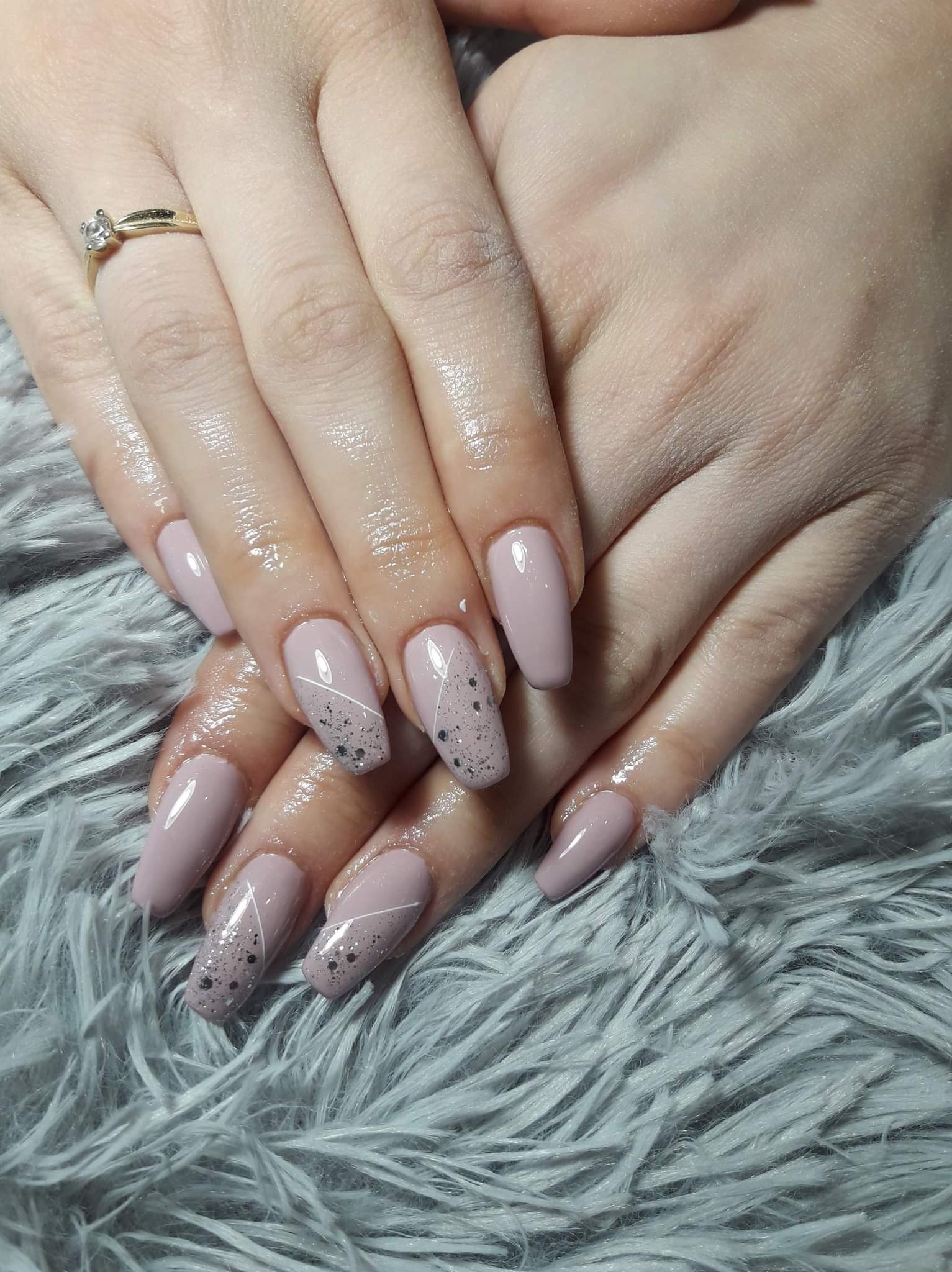 Dłonie z beżowymi, błyszczącymi paznokciami; jeden z paznokci ozdobiony drobnymi czarnymi kropkami i białą linią; na palcu złoty pierścionek z oczkiem; tło stanowi puszysty materiał.