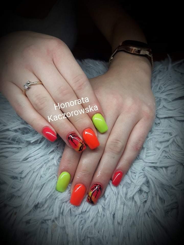 Dłonie z pomalowanymi paznokciami w odcieniach neonowej zieleni, pomarańczy i czerwieni, dwa paznokcie z motywem zachodzącego słońca i palmy, na tle puszystego, jasnoszarego koca.