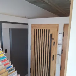 Wystawka z dwoma modelami drzwi: szare drzwi przesuwne z metalową ramą oraz drzwi z jasnego drewna z pionowymi, ciemnymi wstawkami i długim, czarnym uchwytem. W tle surowy, betonowy sufit. Po lewej...