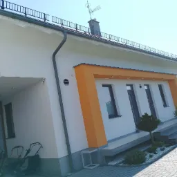 Biała elewacja domu z pomarańczowymi akcentami wokół okien, widoczne trzy kosiarki do trawy pod zadaszeniem.