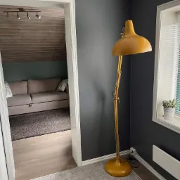 Pokój z szarą ścianą, żółtą lampą podłogową, drewnianym sufitem w oddali i jasną podłogą, po remoncie.