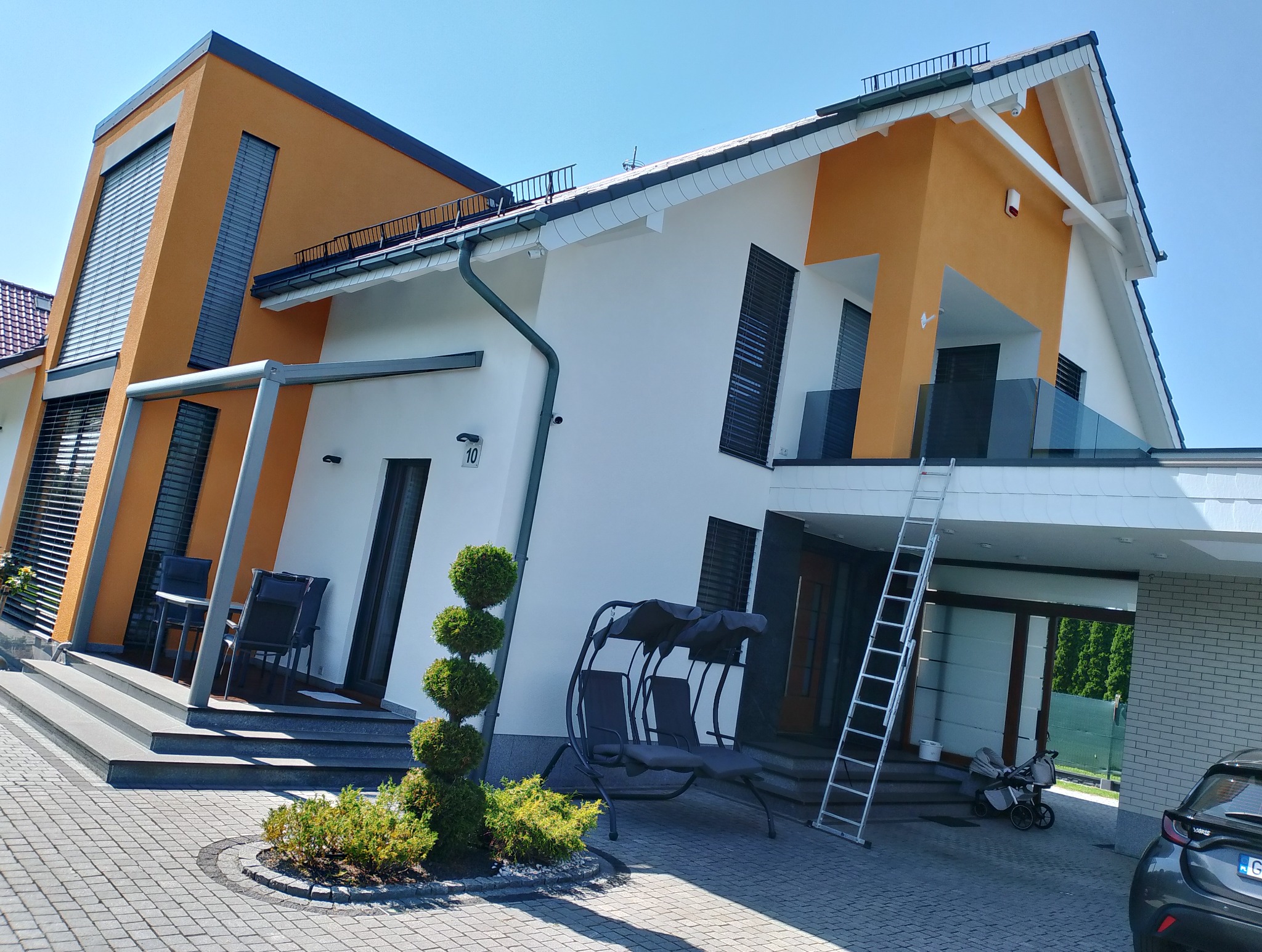 Nowoczesny dom jednorodzinny z pomarańczowo-białą elewacją, na której widać drabinę opartą o balkon, sugerującą prace na wysokościach.