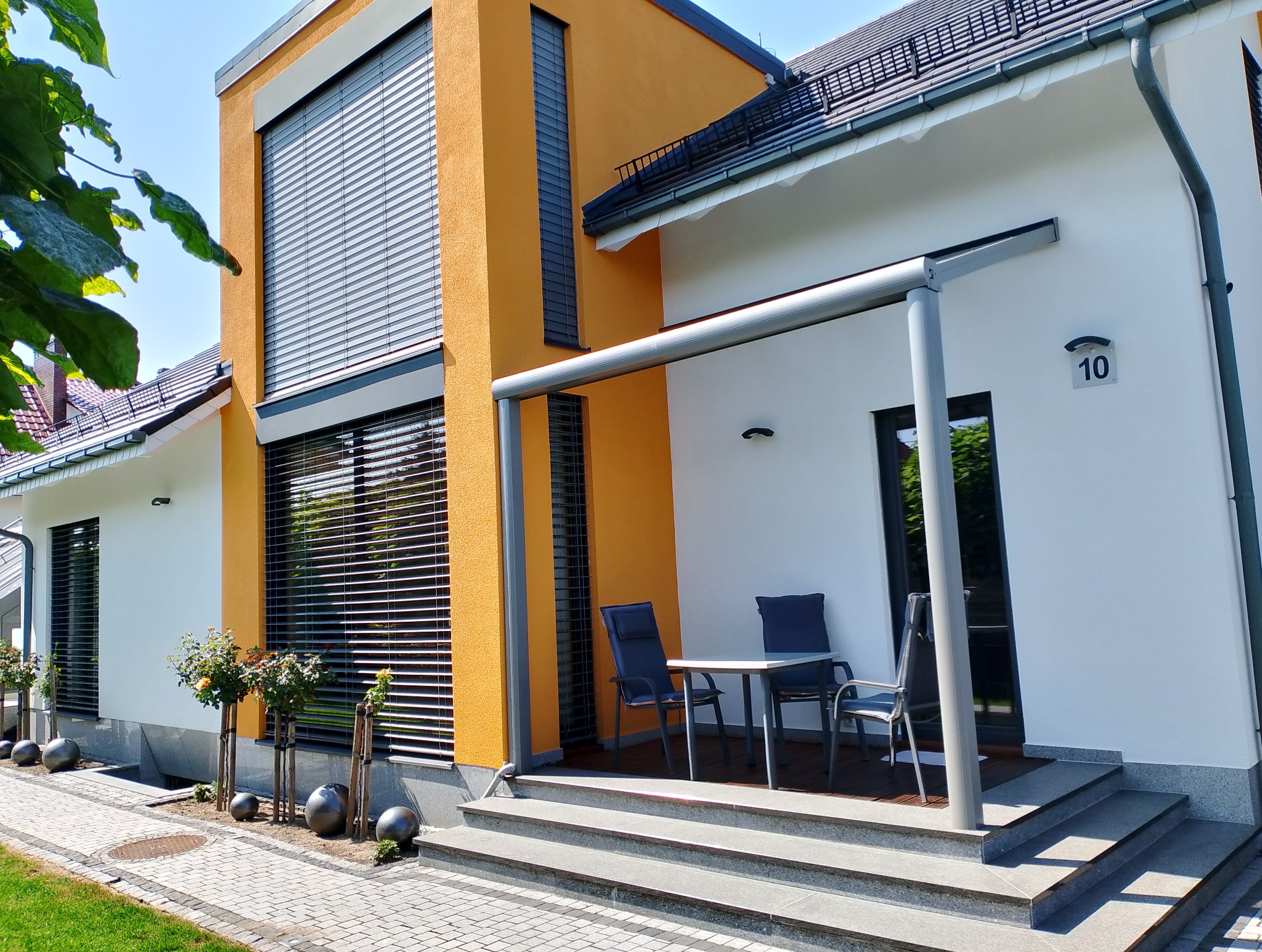 Nowoczesny dom z pomarańczową elewacją, szarymi żaluzjami zewnętrznymi i białymi ścianami, z tarasem i meblami ogrodowymi.