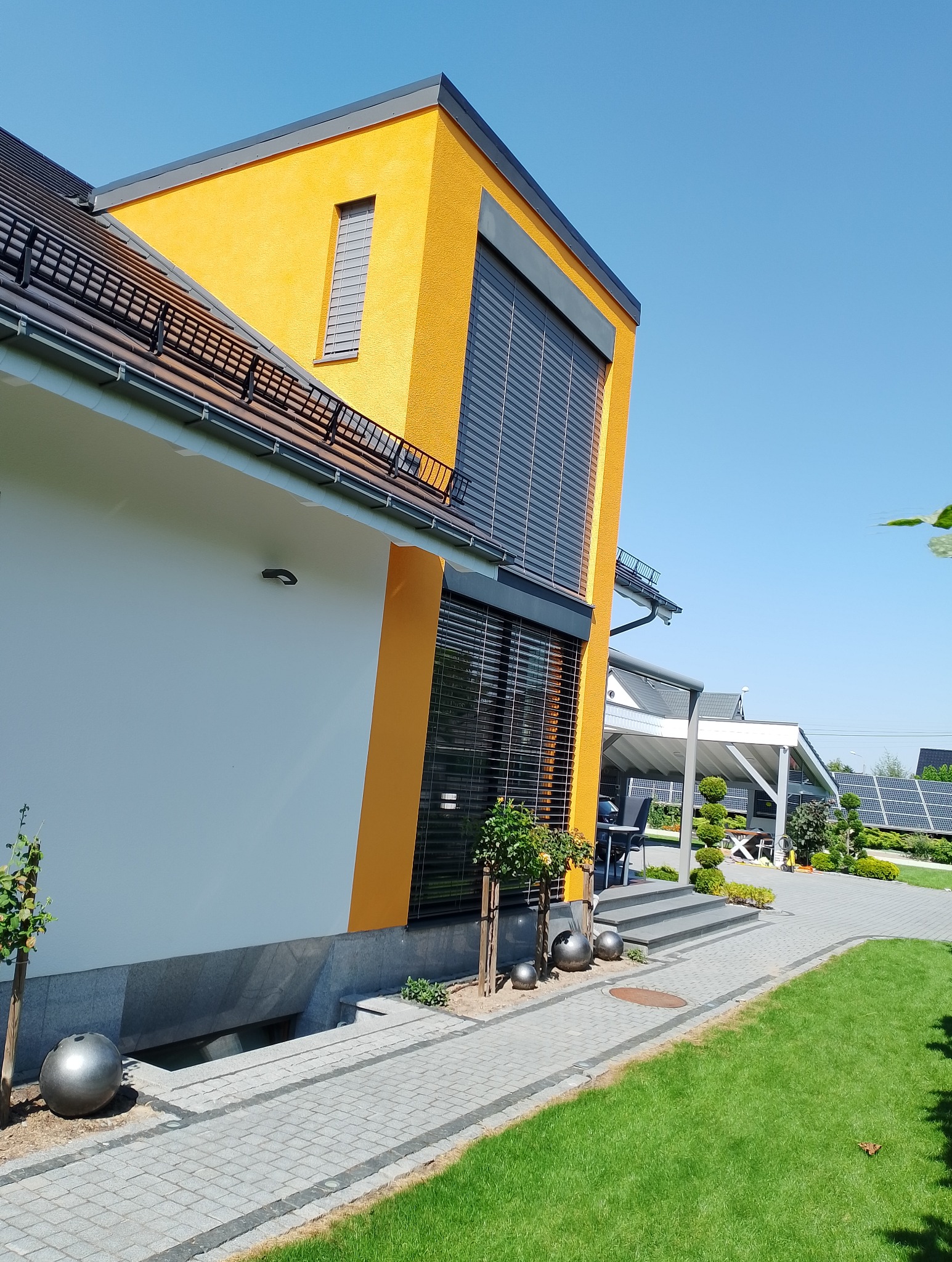 Nowoczesny dom z żółtą elewacją, szarymi roletami i elementami dekoracyjnymi w postaci metalowych kul, widok z poziomu trawnika w słoneczny dzień.