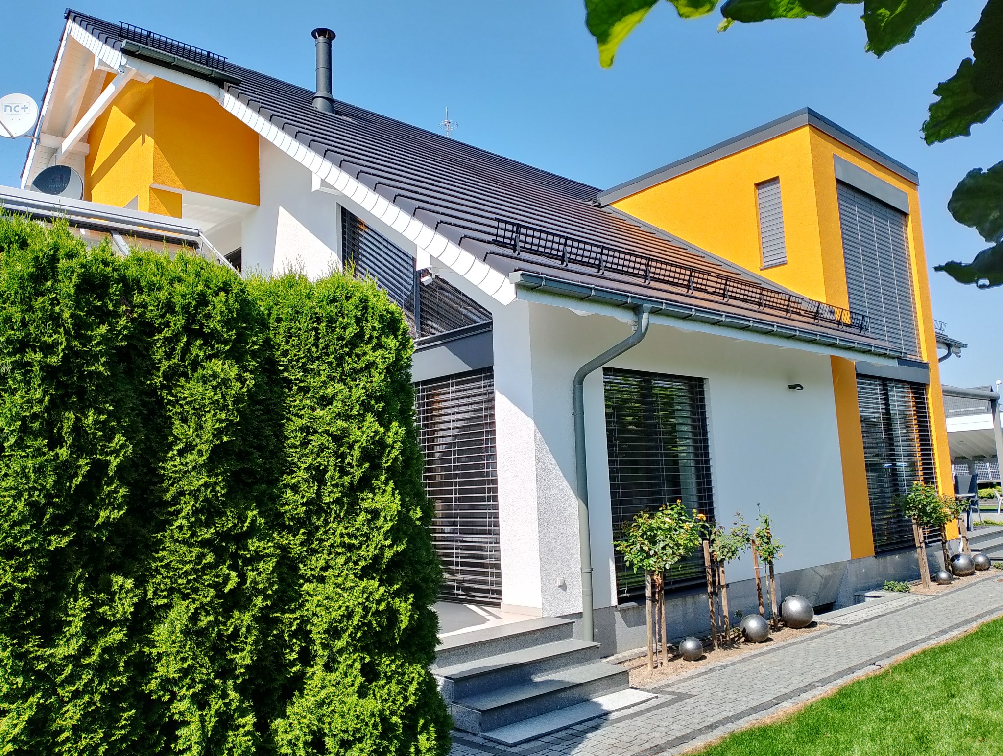 Nowoczesny dom jednorodzinny z żółtą elewacją i szarymi roletami zewnętrznymi, widok z ogrodu z ozdobnymi kulami.