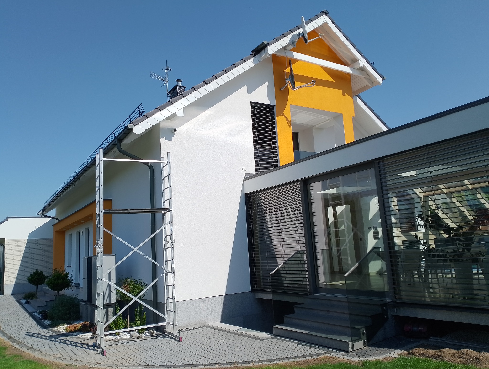Dwukondygnacyjny dom z białą elewacją i pomarańczowymi akcentami, widoczna rozstawiona drabina aluminiowa, błękitne niebo w tle.