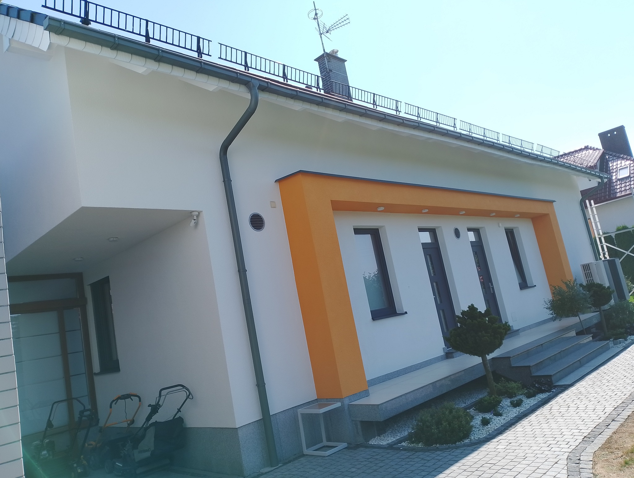 Biała elewacja domu z pomarańczowymi akcentami wokół okien, widoczne trzy kosiarki do trawy pod zadaszeniem.