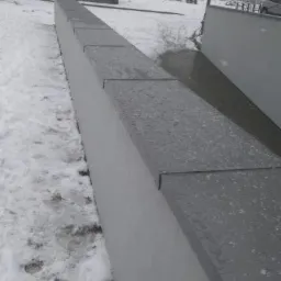 Świeżo wykończona attyka dachu z szarych płyt w zimowej scenerii, widoczne płatki śniegu, w tle budynki mieszkalne.