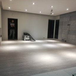 Usługi remontowo- budowlane Grzegorz Siwiec - Jasnoszara podłoga z paneli ułożonych w nowym mieszkaniu, widoczny pracownik w drzwiach i odkurzacz przemysłowy, ściana wyłożona płytkami w odcieniach szarości, sufit z oświetleniem punktowym.