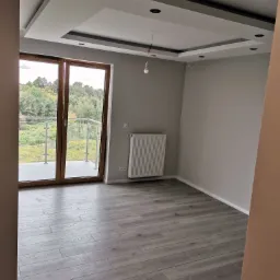 Pusty pokój z szarą podłogą imitującą drewno, przeszklonymi drzwiami balkonowymi z widokiem na drzewa, białym grzejnikiem i dekoracyjnym sufitem podwieszanym z punktowym oświetleniem, oczekujący...