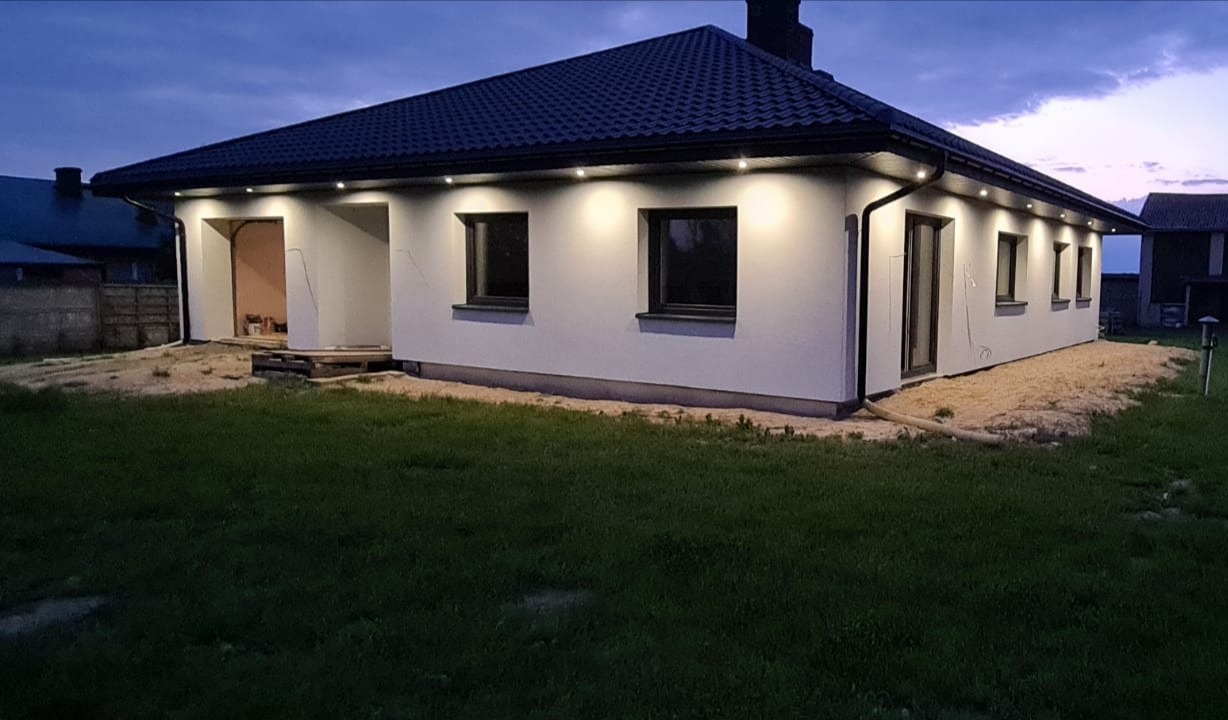 Oświetlony dom jednorodzinny z białym tynkiem zewnętrznym i ciemną dachówką, widoczne okna z ciemnymi ramami i oświetlenie punktowe pod dachem, teren wokół domu częściowo przygotowany pod dalsze...