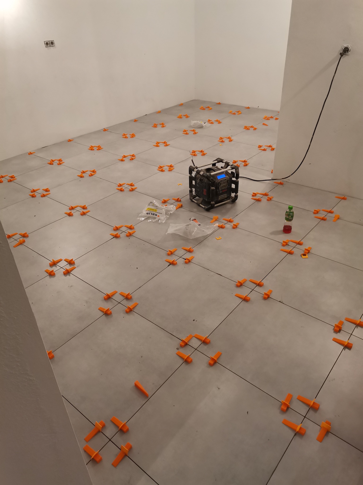 Szare płytki podłogowe z pomarańczowymi klipsami poziomującymi, paczka klipsów SOLID oraz poziomica laserowa na środku pomieszczenia podczas układania płytek.