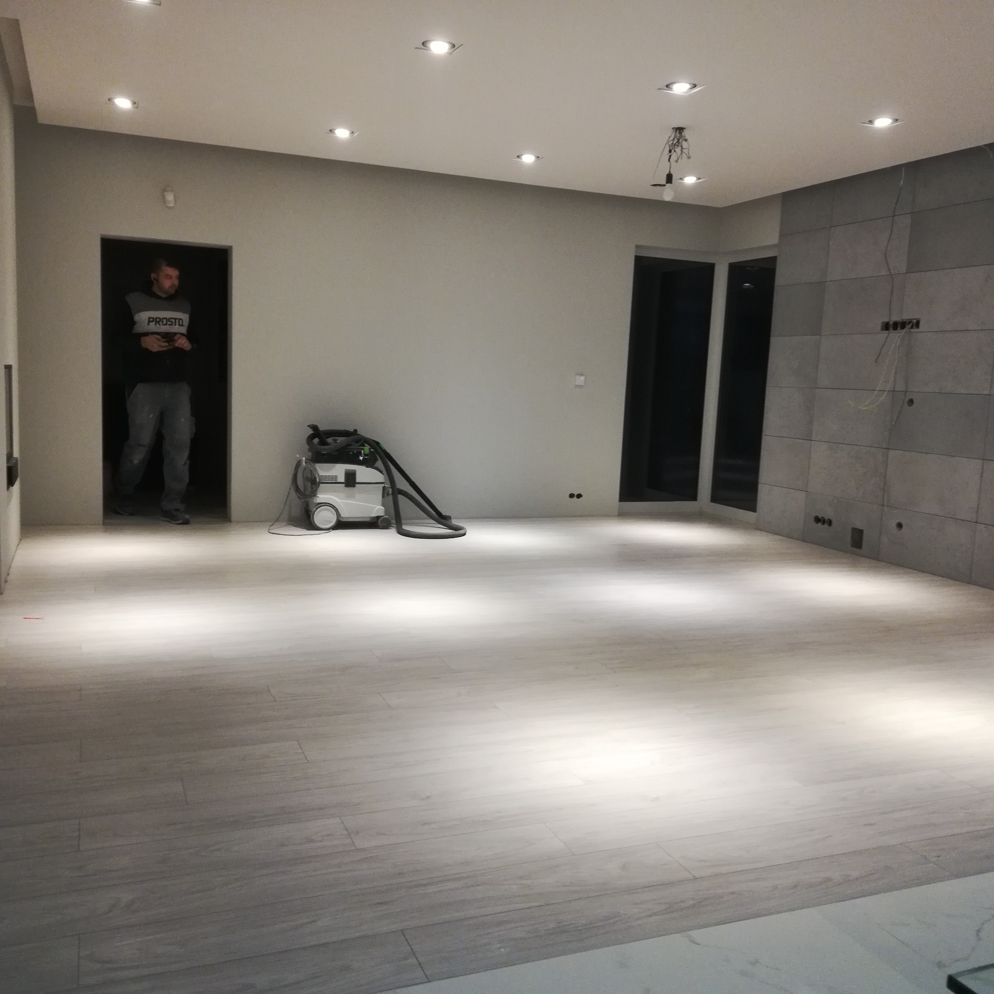 Jasnoszara podłoga z paneli ułożonych w nowym mieszkaniu, widoczny pracownik w drzwiach i odkurzacz przemysłowy, ściana wyłożona płytkami w odcieniach szarości, sufit z oświetleniem punktowym.