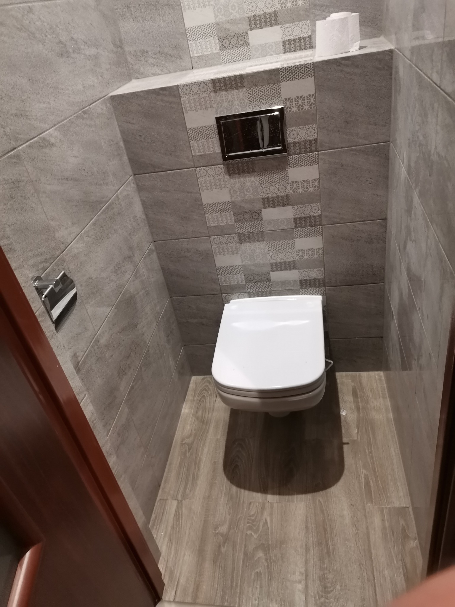 Wnętrze małej toalety z wiszącą muszlą klozetową, szarą glazurą na ścianach i podłodze imitującą drewno, widok zza uchylonych, ciemnych drzwi.