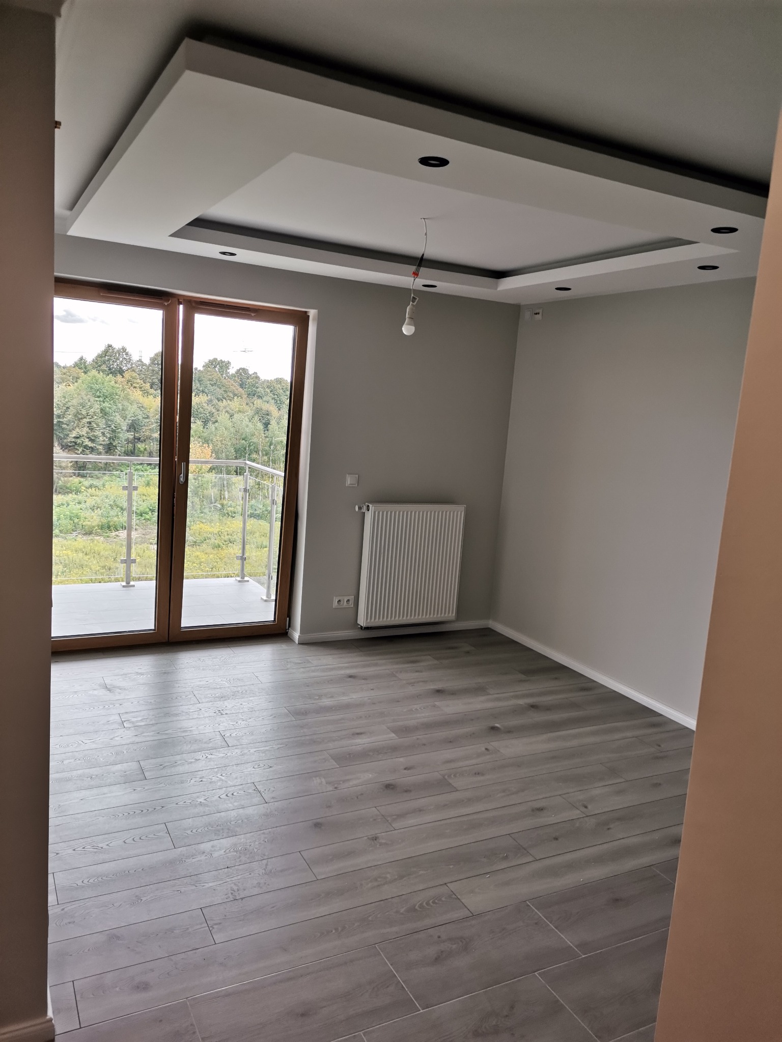 Pusty pokój z szarą podłogą imitującą drewno, przeszklonymi drzwiami balkonowymi z widokiem na drzewa, białym grzejnikiem i dekoracyjnym sufitem podwieszanym z punktowym oświetleniem, oczekujący...