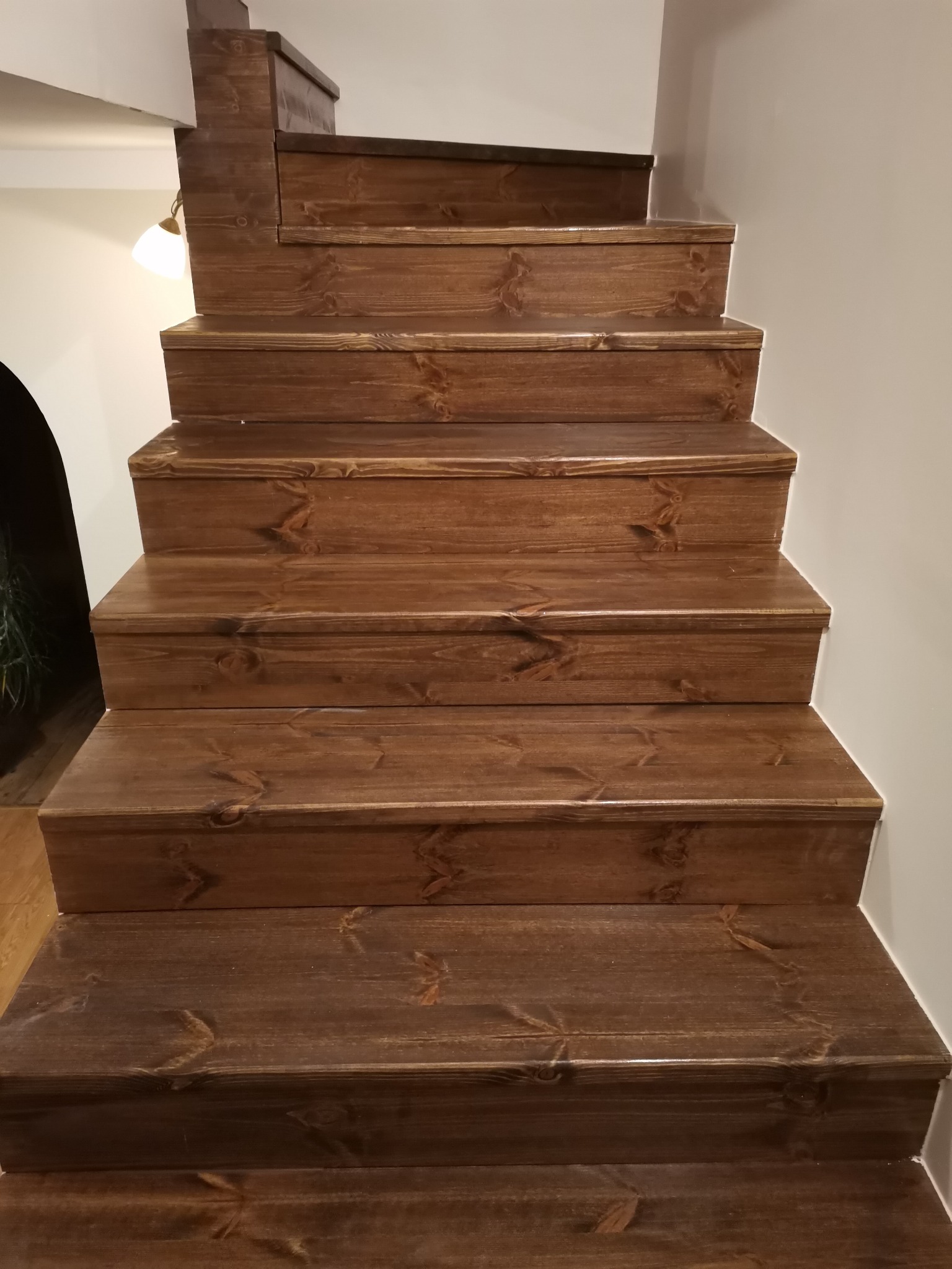 Drewniane schody z ciemnego drewna, widok z dołu, z widocznym oświetleniem w tle i fragmentem ściany.