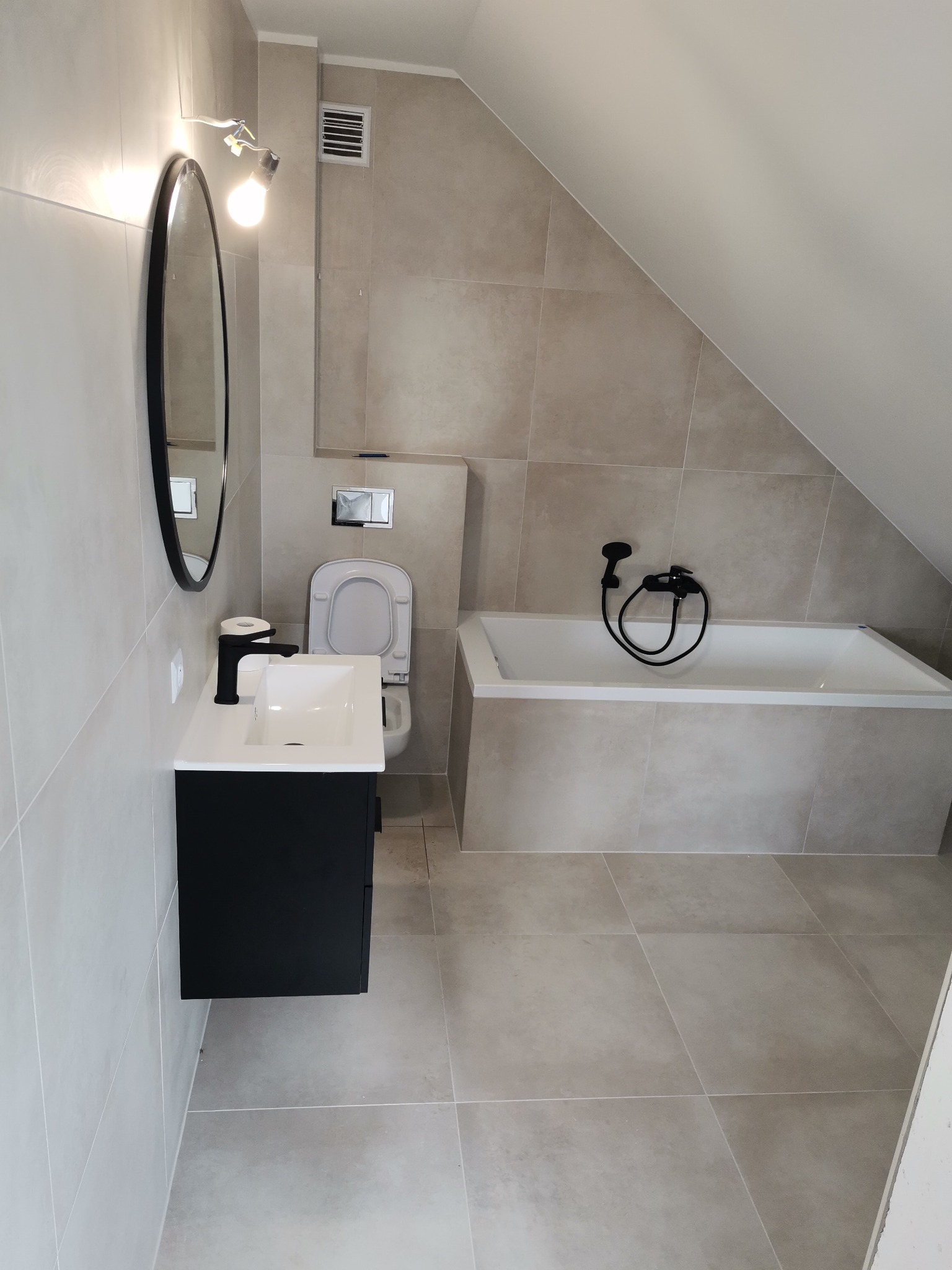 Wykończona łazienka na poddaszu ze skośnym sufitem, beżowe płytki na ścianach i podłodze, czarna bateria umywalkowa i owalne lustro, biała wanna z czarną słuchawką prysznicową.