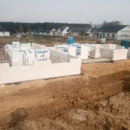 Wznoszenie ścian parteru domu z betonu komórkowego, widoczne zbrojenia, na paletach zapakowane bloczki Solbet, w tle zabudowa jednorodzinna i błękitny samochód dostawczy.