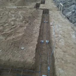 Ziemny wykop z ułożonym zbrojeniem stalowym w przygotowaniu do wylania fundamentów pod budynek, widoczne betonowe podkładki dystansowe.