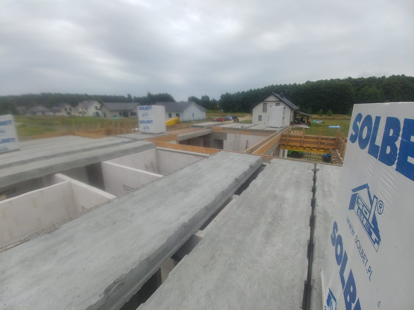 Budowa domu w stanie surowym otwartym: widok z góry na strop z prefabrykatów betonowych, ściany fundamentowe i szalunki drewniane, w tle zabudowa jednorodzinna i las, na pierwszym planie opakowanie...