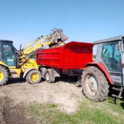 Żółta koparko-ładowarka marki Hidromek nabiera ziemię i wysypuje ją do czerwonej przyczepy ciągniętej przez szaro-czerwony traktor Autotronic, widoczne ślady opon na piaszczystym podłożu i błękitne...