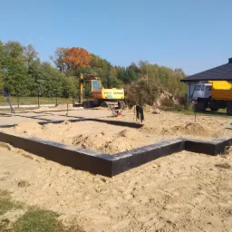 Przygotowany teren pod budowę domu jednorodzinnego: wytyczone i zabezpieczone fundamenty, żółty koparko-ładowarka Zeppelin oraz wywrotka w tle, jesienne drzewa z pomarańczowymi liśćmi.
