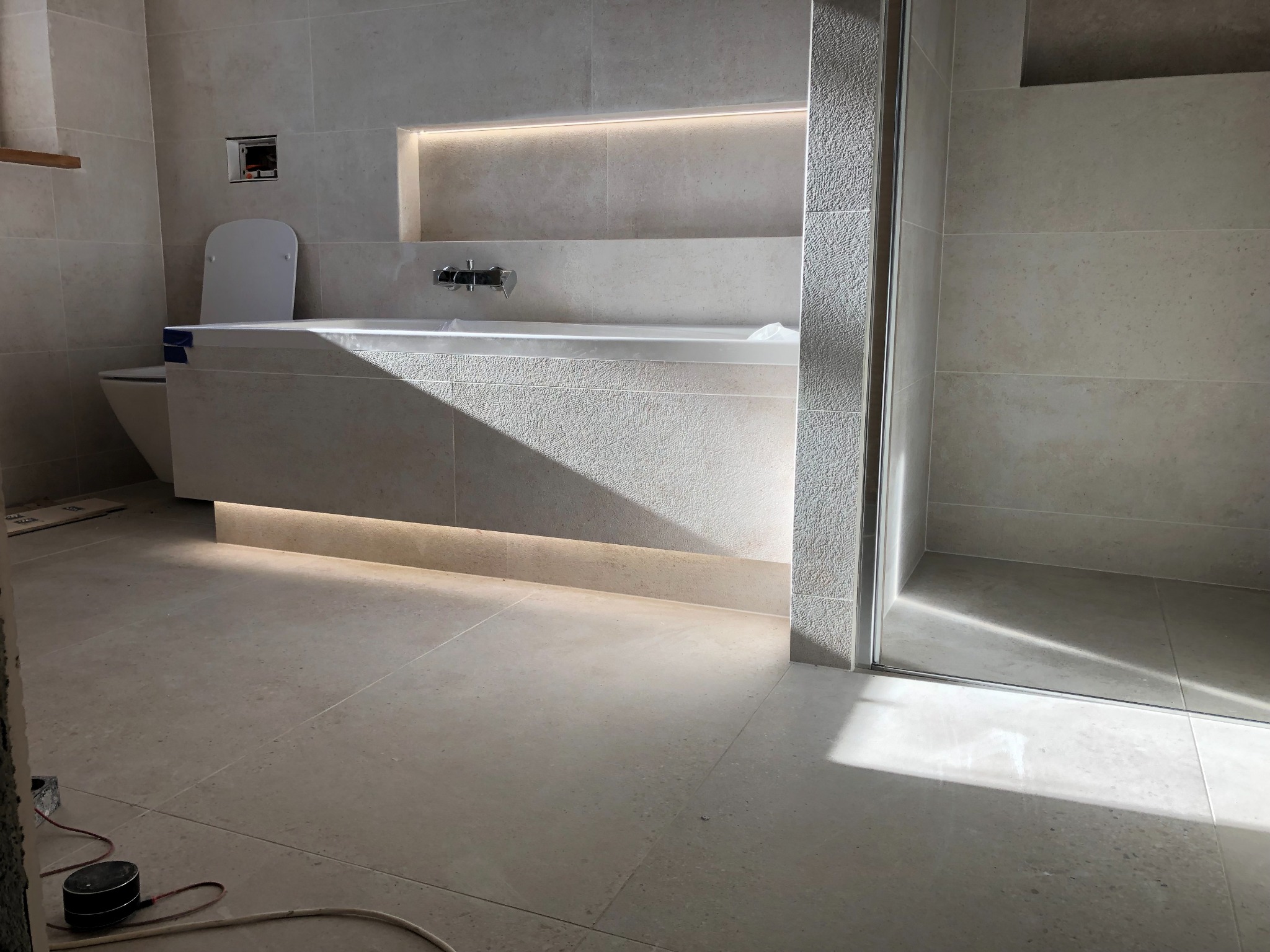 Nowoczesna łazienka w jasnych odcieniach beżu z podświetlaną wnęką nad wanną, widoczna toaleta i kabina prysznicowa, minimalistyczny design.