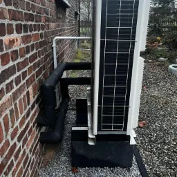 Zewnętrzna jednostka pompy ciepła zamontowana przy ceglanej ścianie budynku, z widocznymi rurami izolowanymi termicznie i podstawą z czarnych bloczków na żwirowym podłożu.