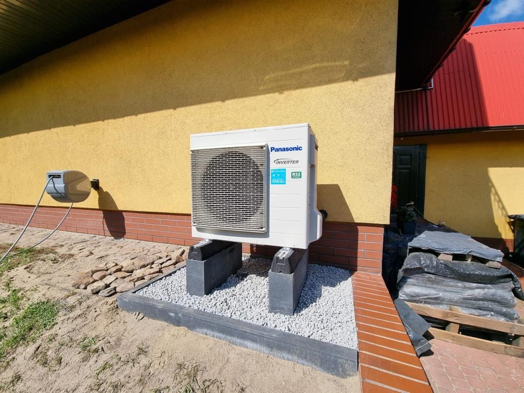 Zewnętrzna jednostka klimatyzacji Panasonic Inverter, zamontowana na betonowych podstawach z szarym żwirem, przy żółtej ścianie budynku z czerwonym dachem, widoczny zwinięty kabel na ścianie.