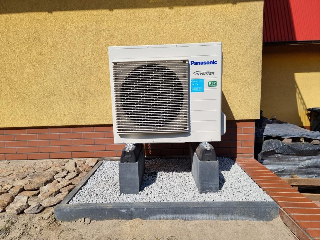 Zewnętrzna jednostka klimatyzacji Panasonic Inverter R32 umieszczona na betonowych podstawach, z widoczną elewacją budynku w kolorze żółtym i obramowaniem z kamieni.