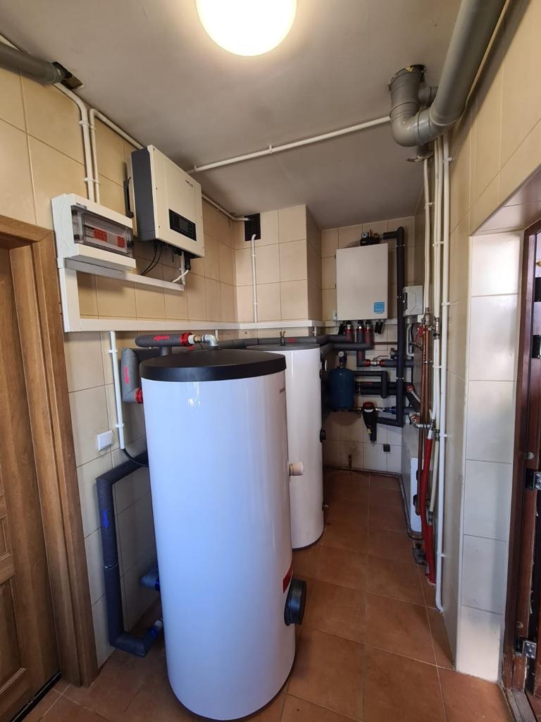 Dwa białe zasobniki ciepłej wody użytkowej w kotłowni z beżowymi płytkami, widoczne rury, zawory, skrzynka elektryczna i falownik na ścianie.