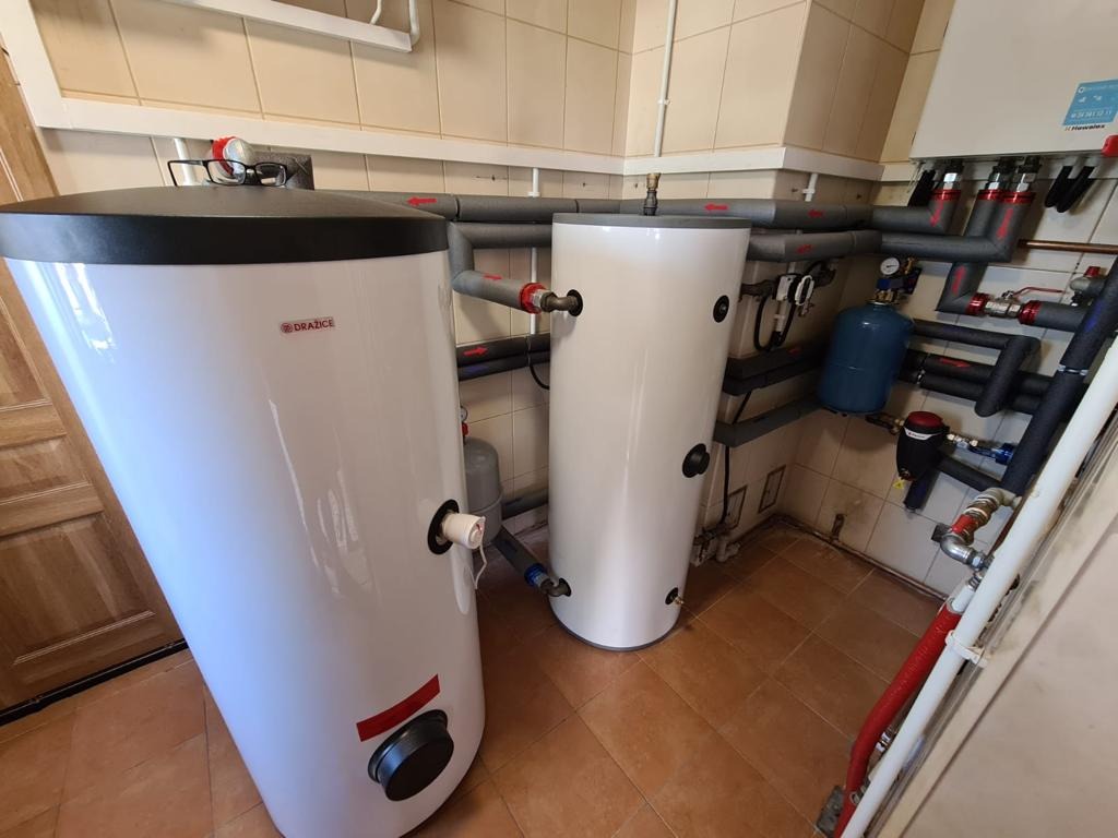 Dwa białe, cylindryczne zbiorniki akumulacyjne ciepła marki Drazice w kotłowni, z widocznymi rurami instalacji grzewczej i osprzętem, na tle beżowych płytek ściennych i brązowej podłogi.