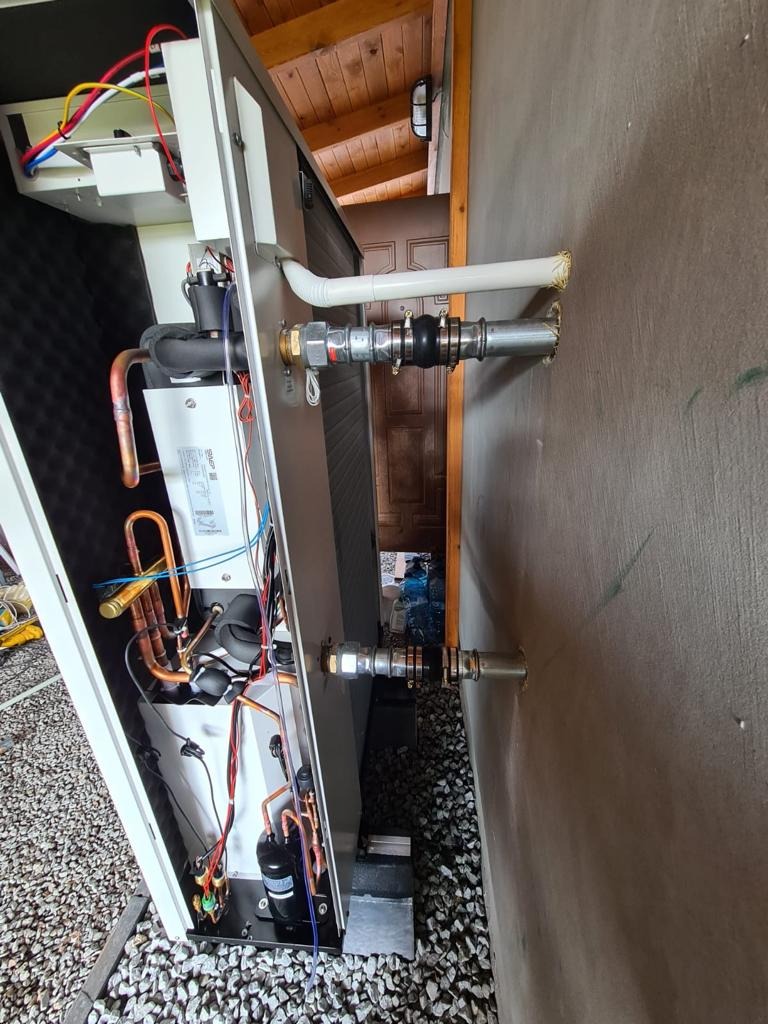 Pompa ciepła powietrze-woda w trakcie instalacji, widoczne podłączenia hydrauliczne do ściany budynku, elementy instalacji elektrycznej i chłodniczej, oraz otoczenie z kamieniami.