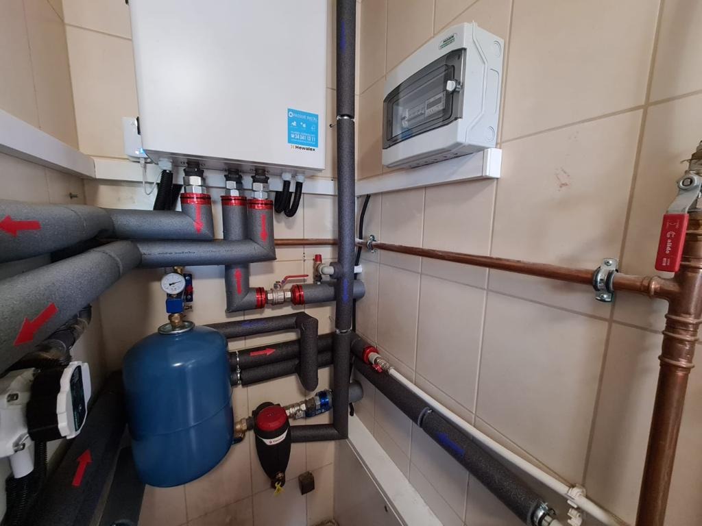 Instalacja grzewcza z elementami pompy ciepła: rury w izolacji z czerwonymi strzałkami, manometr, niebieski zbiornik wyrównawczy, biała skrzynka sterownicza Hewalex, kran z czerwoną rączką, na tle...
