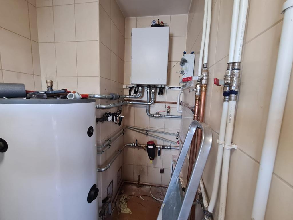 Wnętrze kotłowni z zamontowanym białym zasobnikiem ciepłej wody i kotłem grzewczym na ścianie, widoczne rury instalacyjne, zawory, filtr oraz drabina oparta o ścianę.