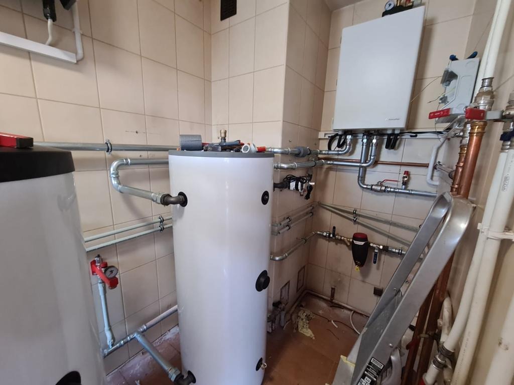 Instalacja z dwoma białymi zasobnikami ciepłej wody, rurami miedzianymi i aluminiową drabiną w pomieszczeniu wyłożonym kafelkami.
