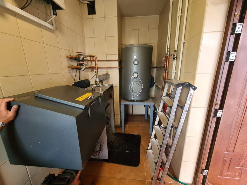 Instalacja nowego zbiornika buforowego obok istniejącego pieca w kotłowni z beżowymi płytkami; widoczna drabina oparta o ścianę i miedziane rury instalacji grzewczej.