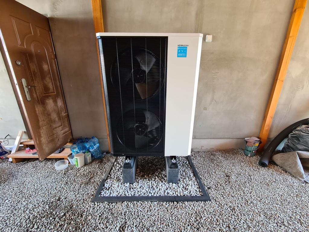Zewnętrzna pompa ciepła marki Hewalex, ustawiona na betonowych blokach na żwirowym podłożu, widoczna otwarta drewniana brama i elementy instalacji w tle.
